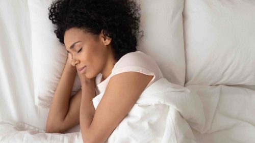 Beautiful African American woman with hands under cheek sleeping in bed under blanket top view, girl with closed eyes resting in bedroom, enjoying fresh bedclothes, lying on soft pillow close up